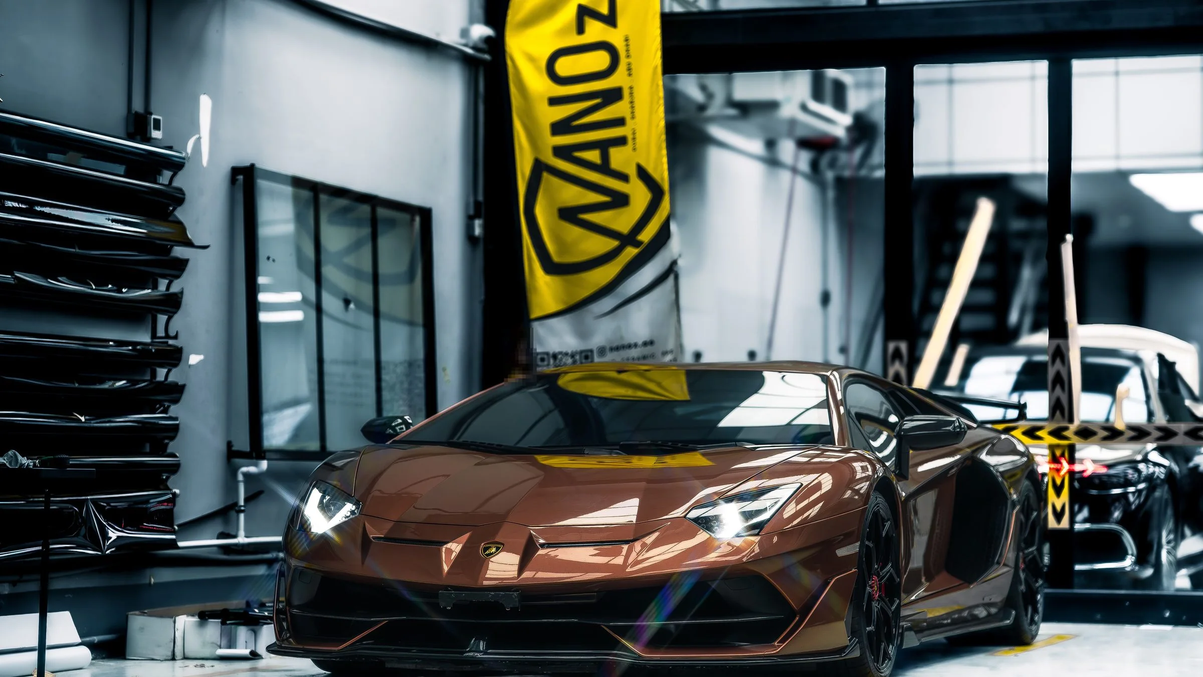 A bronze Lamborghini Aventador SVJ under the roof lights of the Dubai workshop, a gold Nanoz flag behind